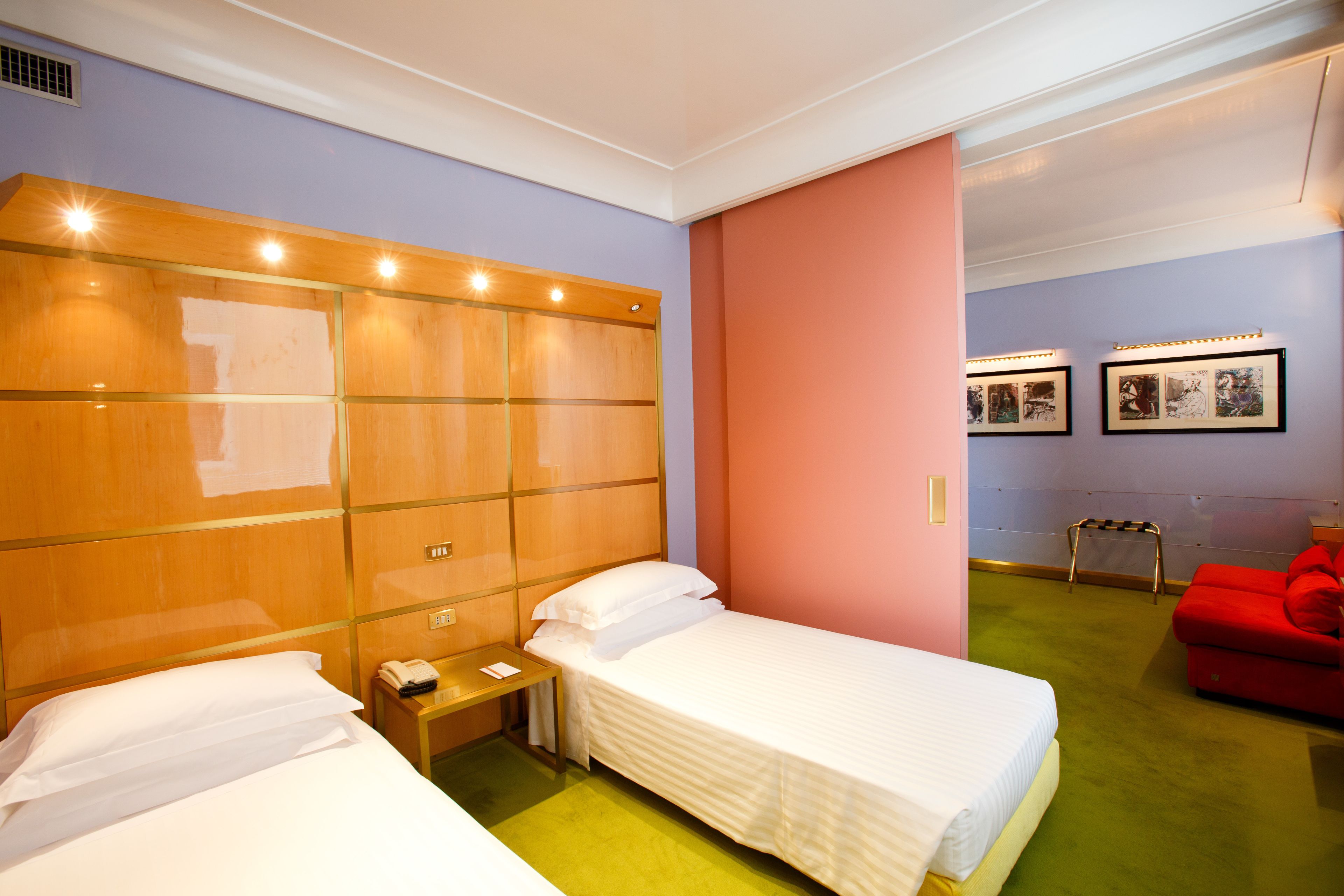 Quarto de hotel com duas camas, uma cabeceira de madeira, uma pequena mesa de cabeceira com telefone e uma cadeira vermelha.