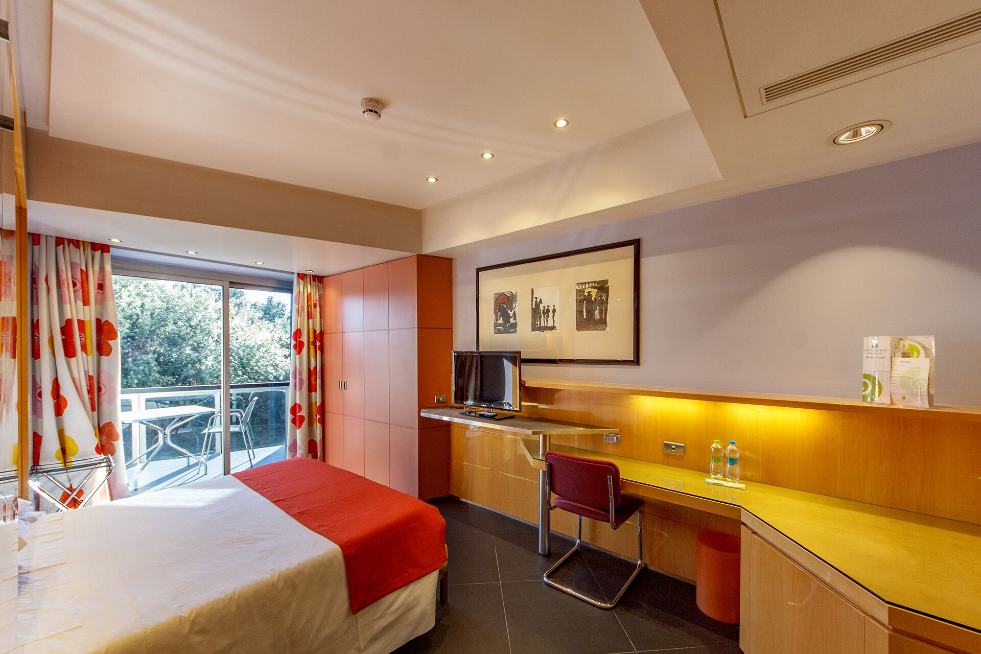A hotel room with a bed, a desk, a chair, a TV, and a balcony with a view of trees.