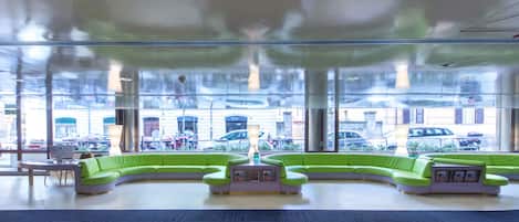 A modern lobby with a curved green sofa area, a reception desk, and large windows offering a view of parked cars outside.