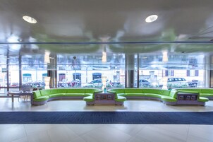Una hall moderna con un'area salotto verde con divano a forma di curva, una reception e ampie finestre con vista sulle auto parcheggiate all'esterno.