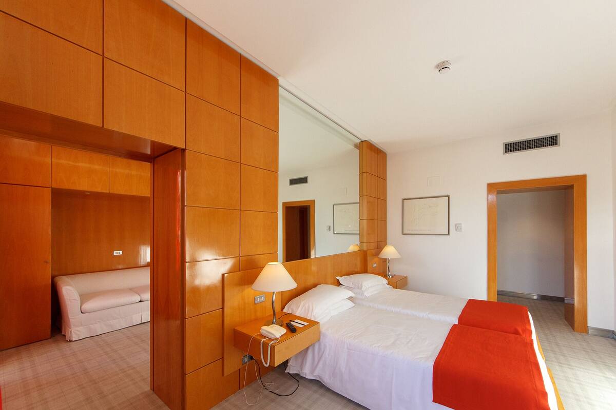 A hotel room with a bed, bedside tables, a mirror, a wardrobe, and a red chair.