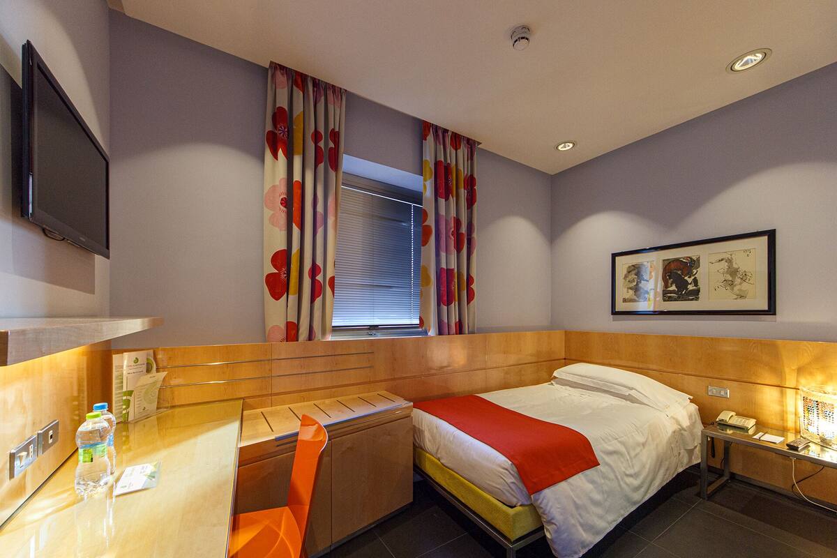 A hotel room with a bed, a desk, a chair, a TV, a window with curtains, and artwork on the wall.