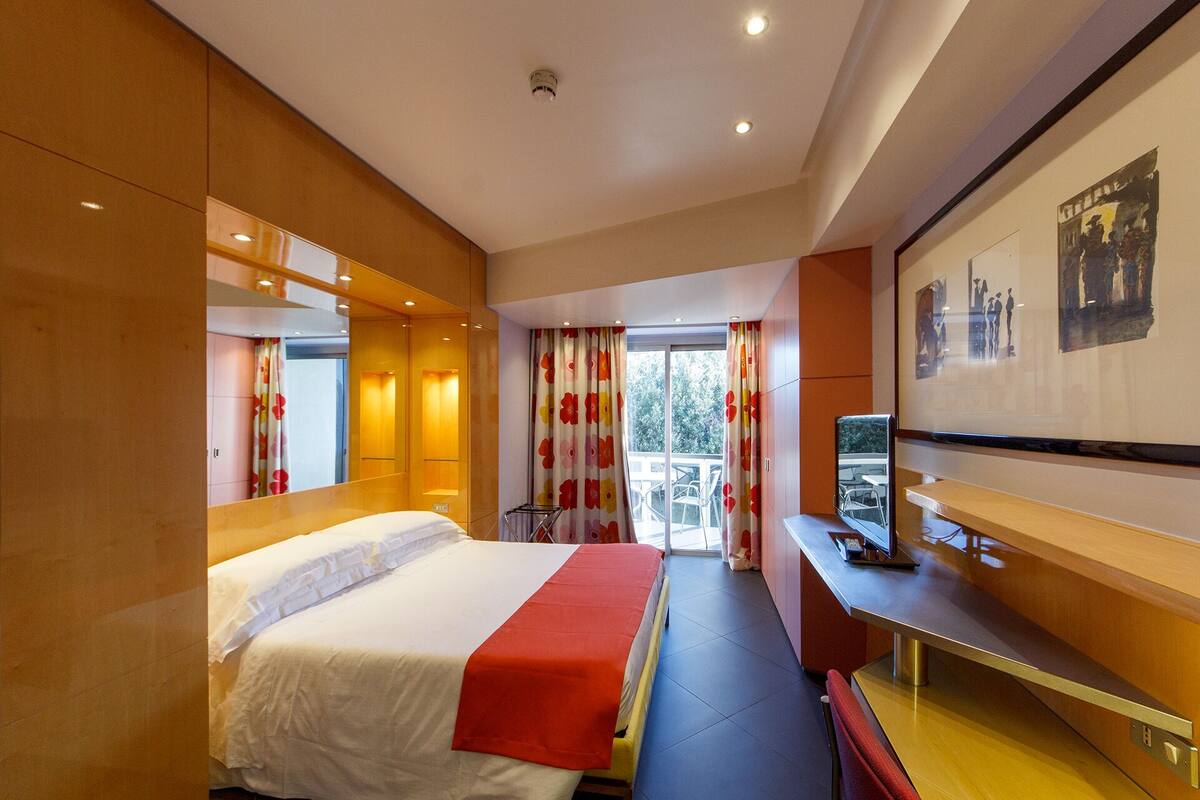 A hotel room with a bed, a desk, a chair, a TV, and a balcony with a view of trees.