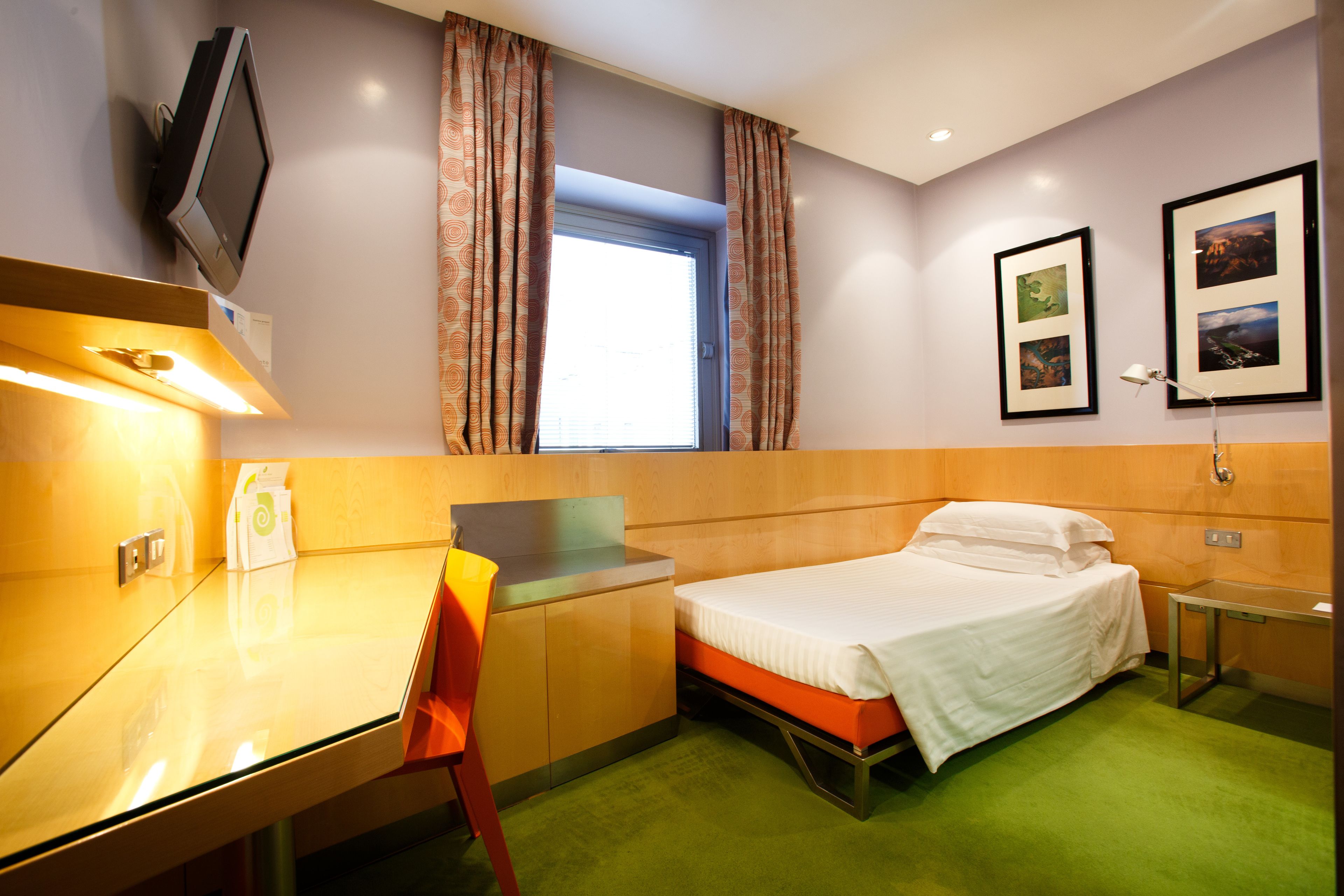 A hotel room with a bed, a desk, a chair, a TV, and two framed pictures on the wall.