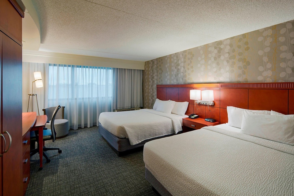 A hotel room with two beds, a desk, a chair, a lamp, and a patterned wall.