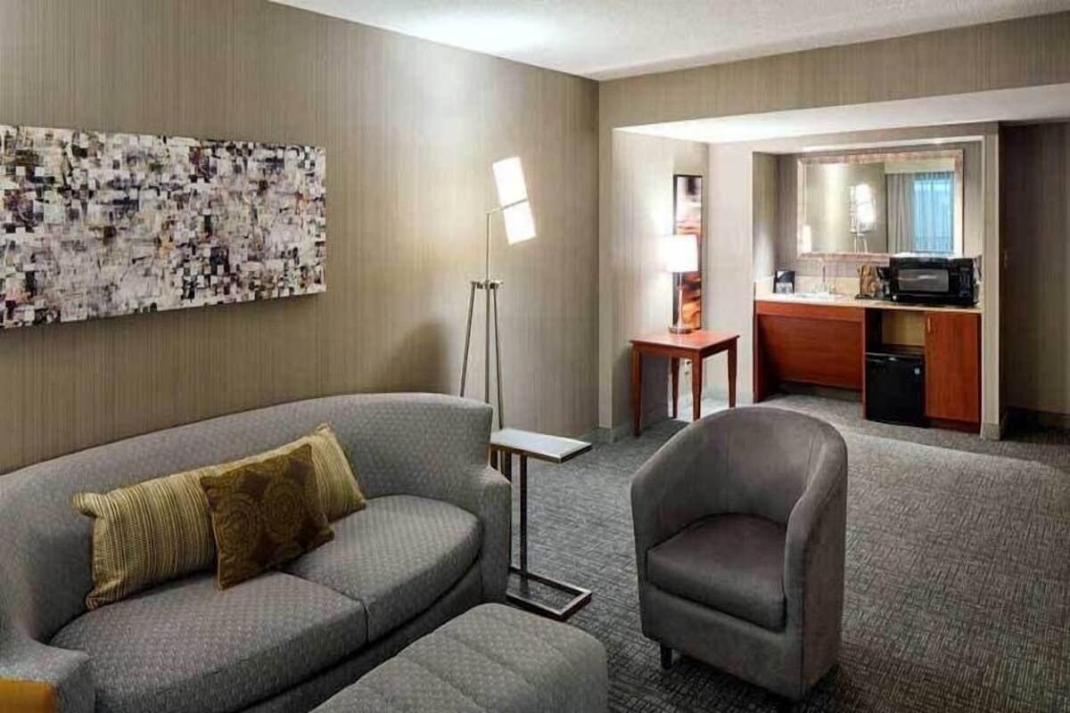 A hotel room with a sofa, armchair, a small table, a lamp, and a kitchenette area.