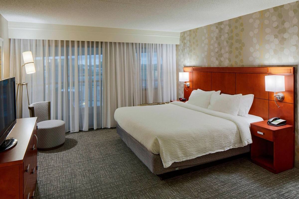 A hotel room with a bed, a desk with a TV, a chair, a nightstand with a phone, and a window with curtains.