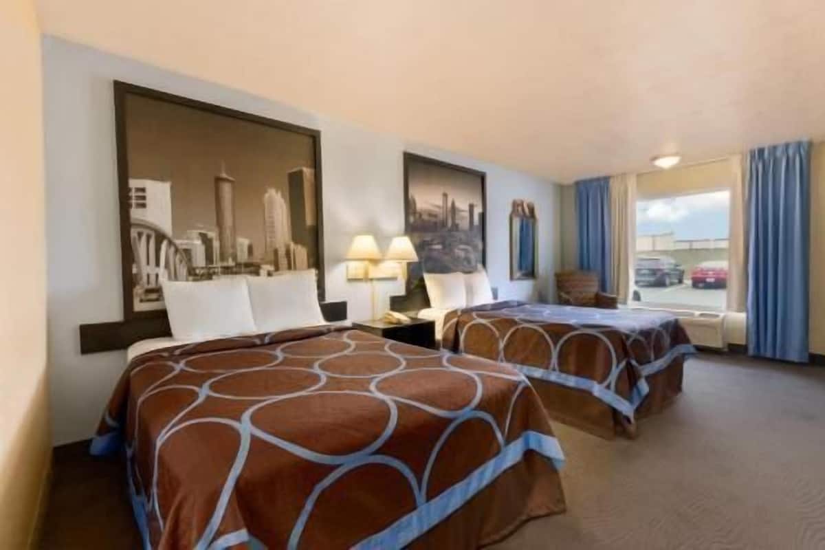 A hotel room with two beds, a cityscape painting, a window with curtains, and a view of parked cars.