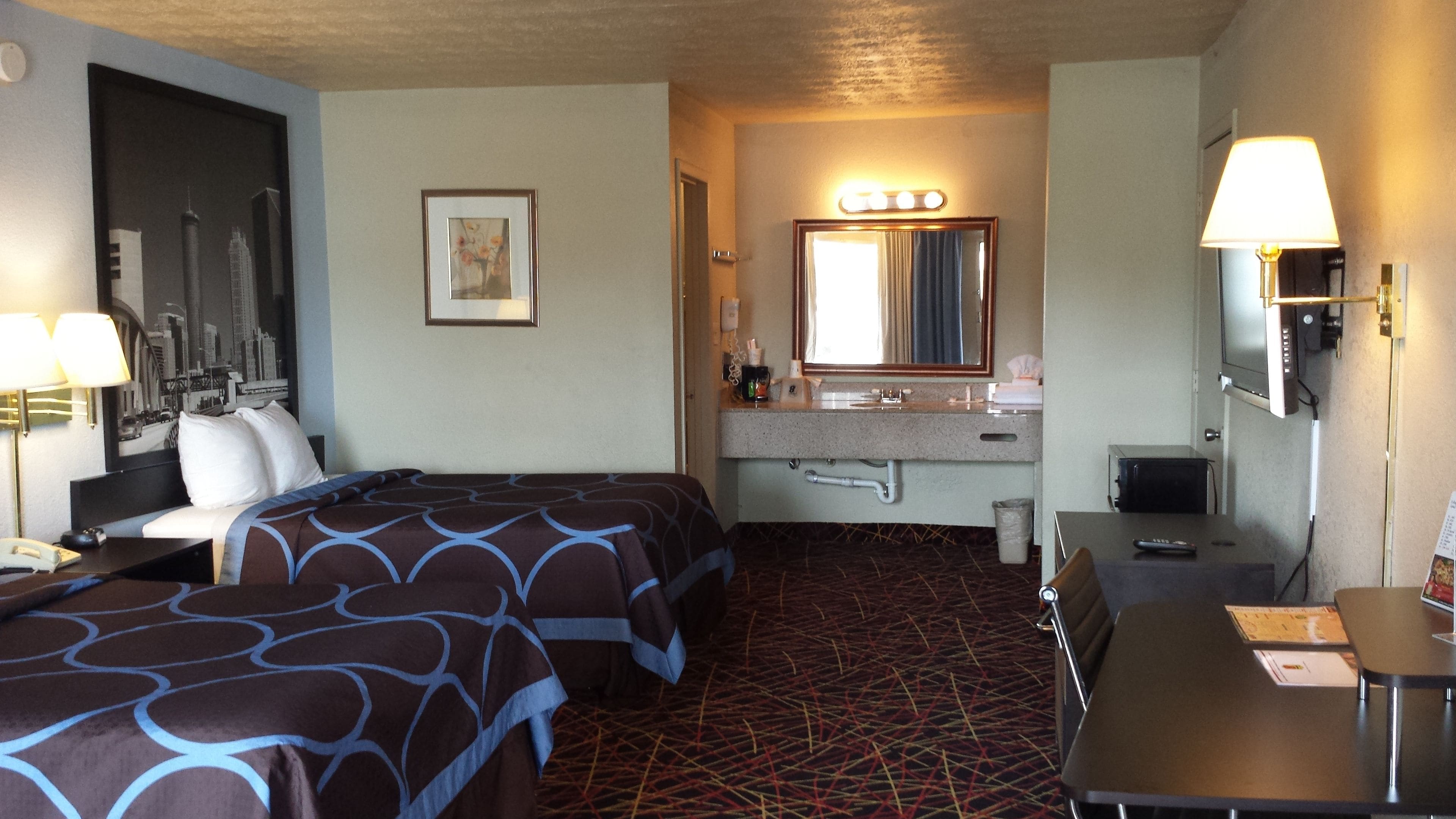 A hotel room with two beds, a bathroom with a sink and mirror, and a desk with a lamp.