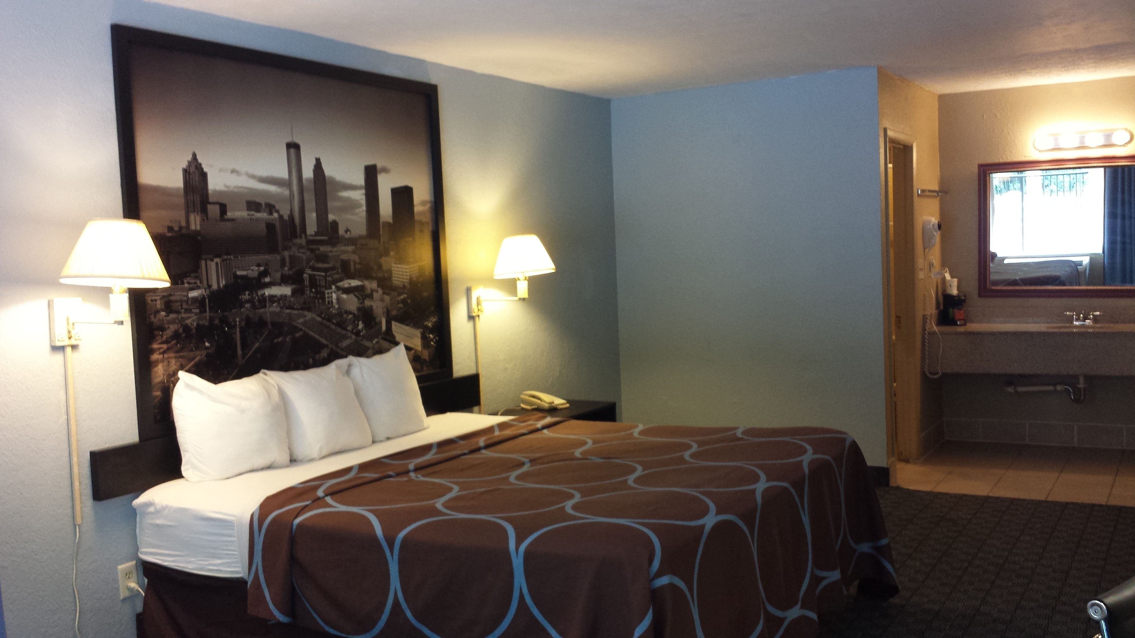 A hotel room with a large bed, two bedside lamps, a cityscape poster on the wall, a bathroom with a sink and mirror, and a carpeted floor.