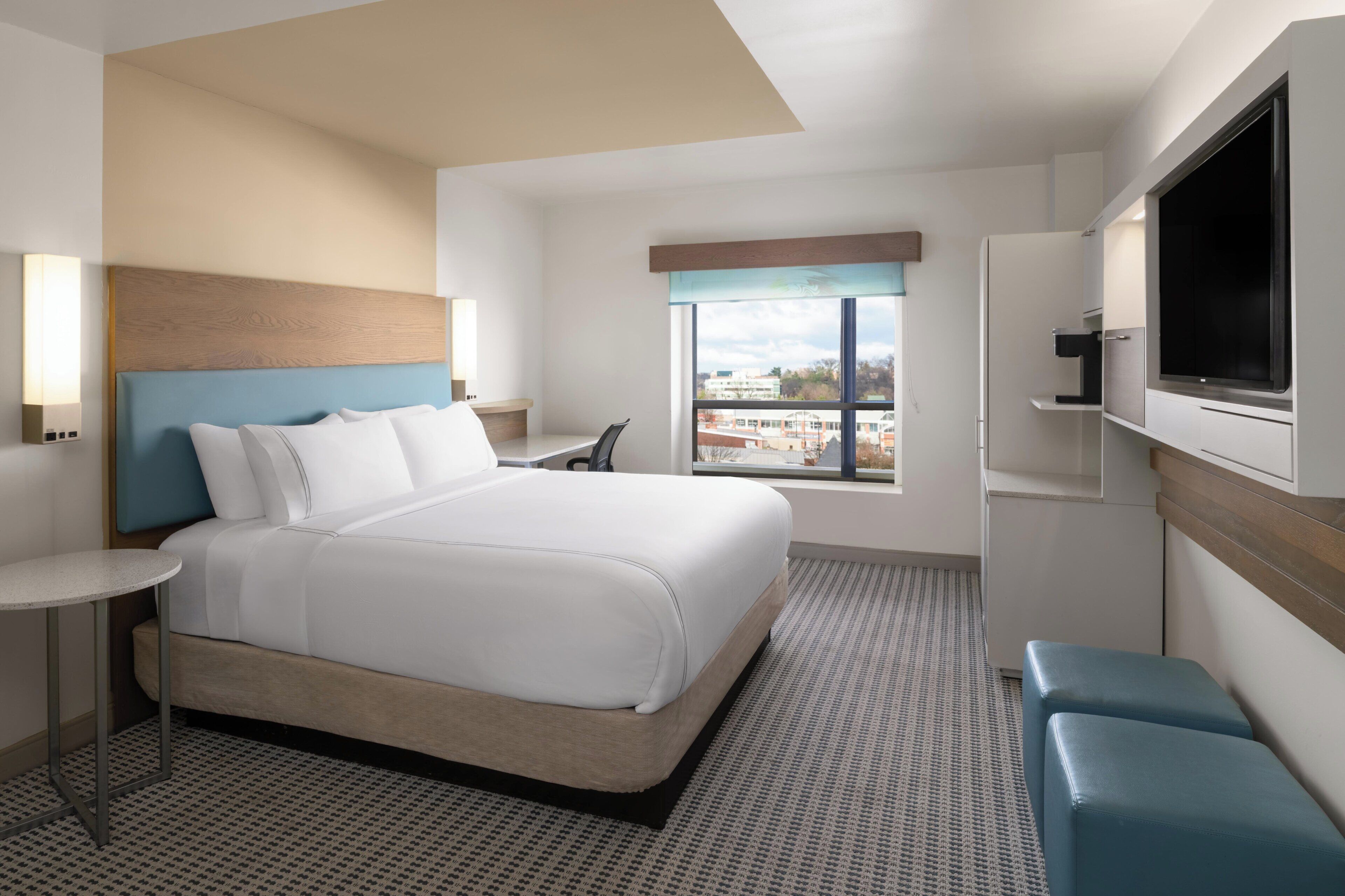 A hotel room with a bed, a desk, a chair, a TV, and a window with a city view.