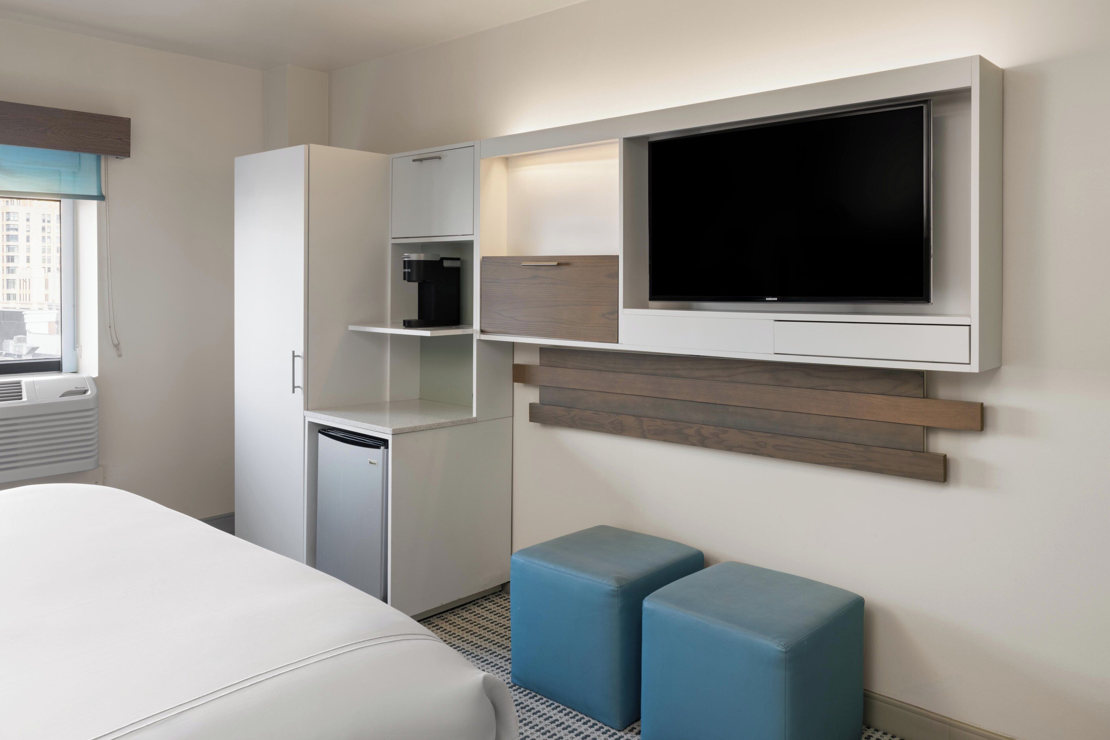 Une chambre d’hôtel équipée d’un lit, d’un téléviseur fixé au mur, d’un mini-réfrigérateur, d’une machine à café et de deux poufs bleus.