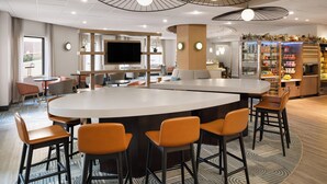 Un hall d’hôtel moderne doté d’un bar central, de différents espaces aménagés pour s’asseoir et d’un téléviseur.