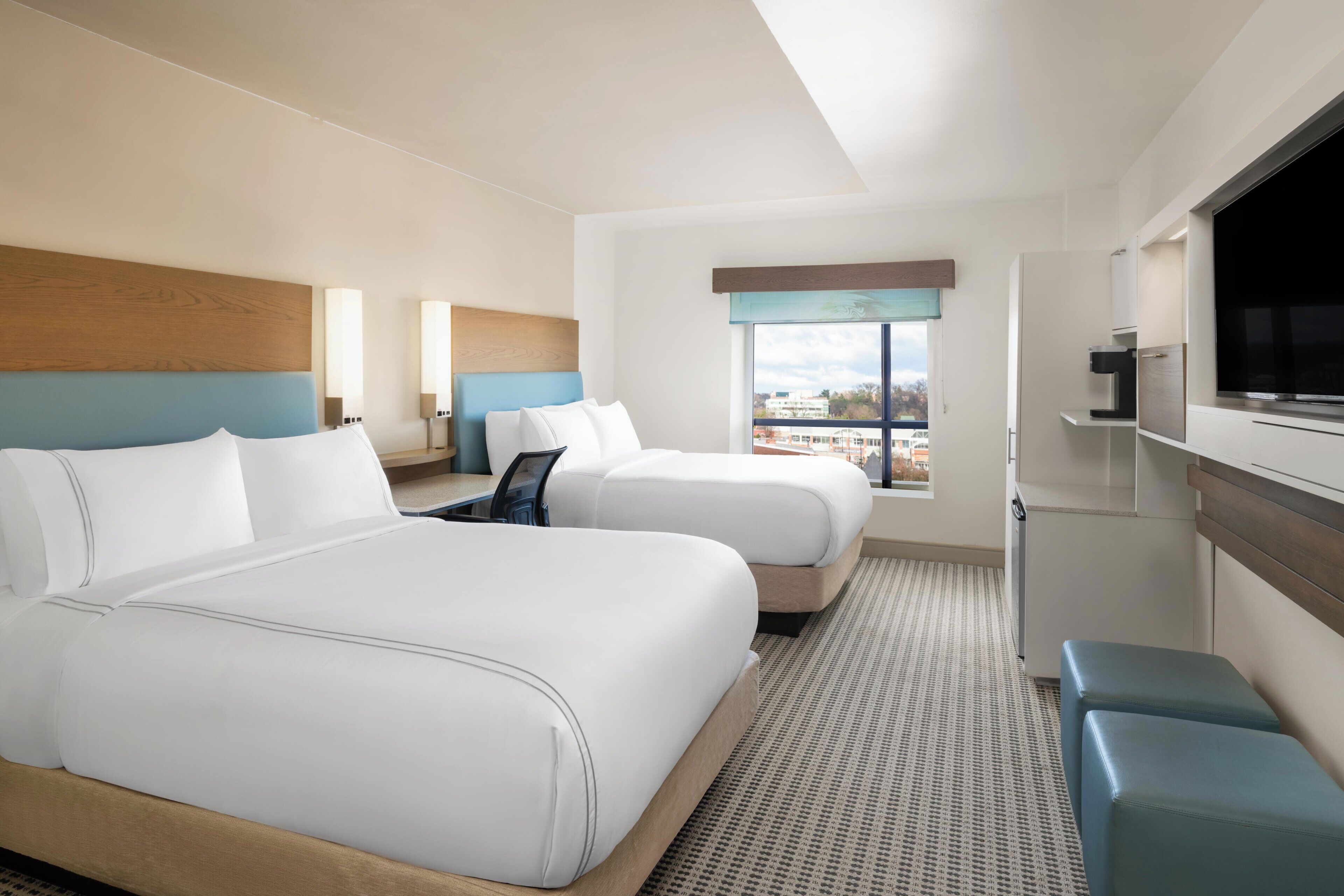 A hotel room with two beds, a TV, a desk, and a window with a city view.