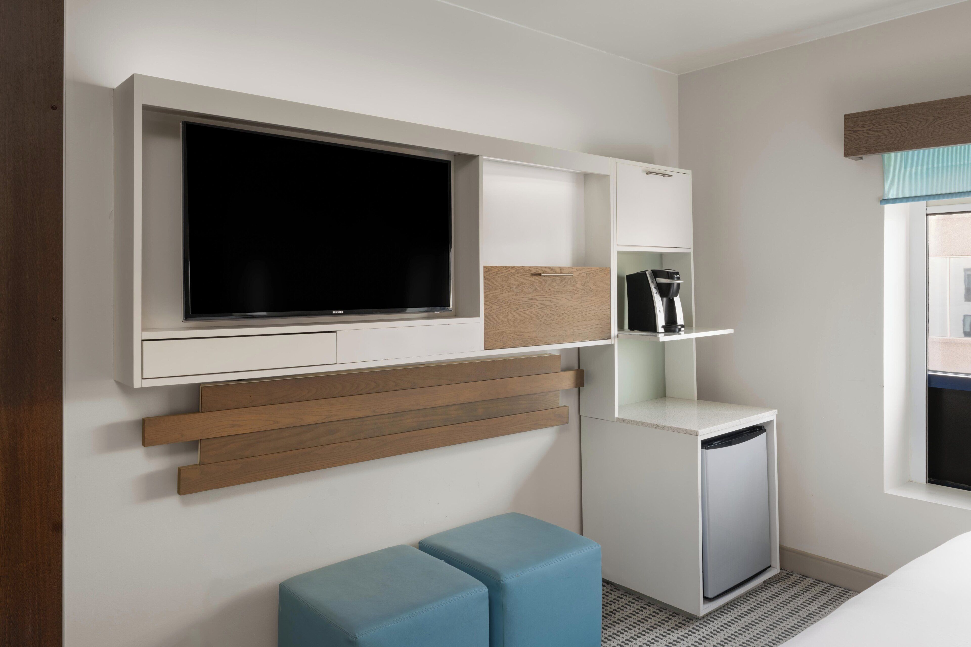 Una habitación de hotel moderna con televisión de pantalla plana, un mini refrigerador, cafetera y un pequeño rincón de descanso.