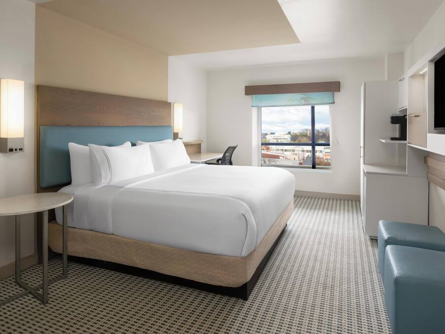 Quarto de hotel moderno com uma cama grande, uma mesinha, uma televisão e uma janela com vista para a cidade.