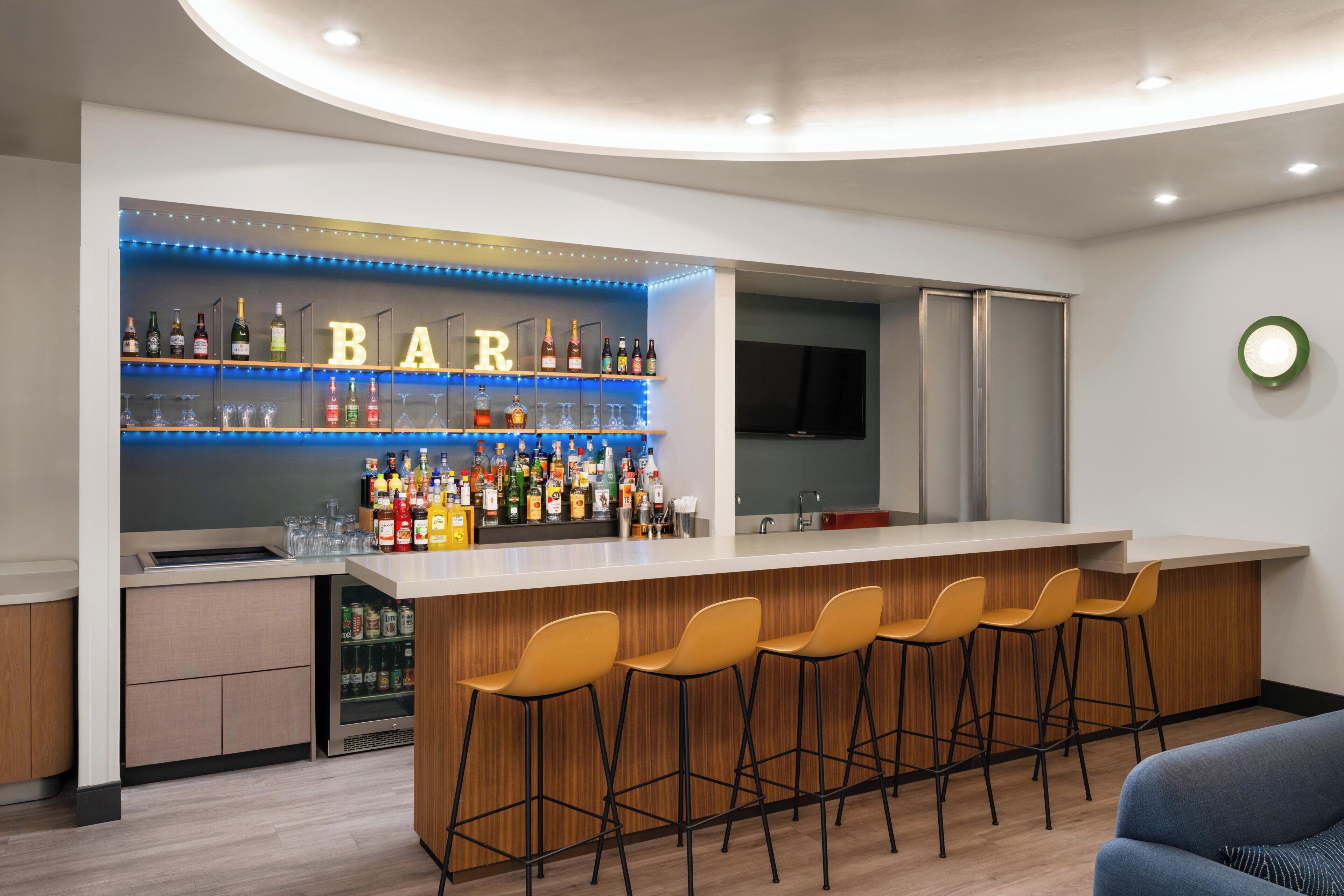 Un área de bar moderna con mostrador, taburetes, estantes con botellas y un televisor.