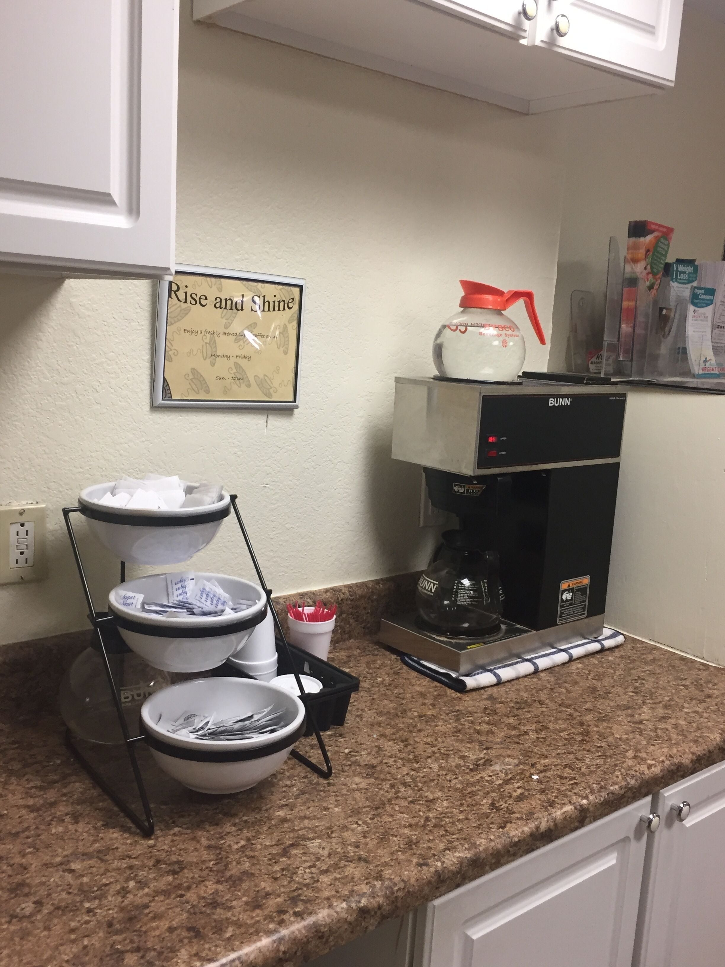 A kitchen with a coffee maker, a coffee dispenser, and a sign that says "Rise and Shine".