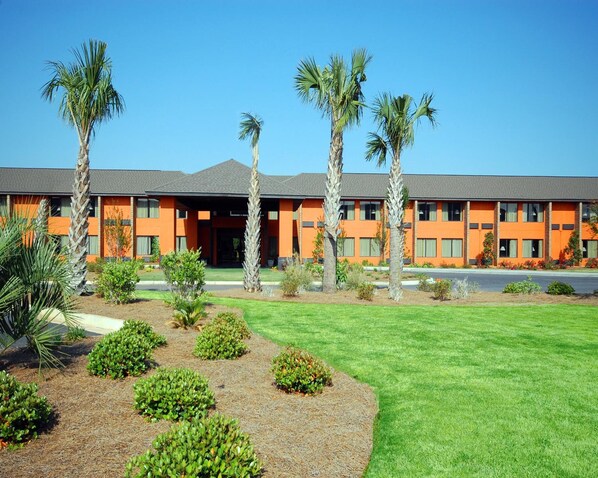 A hotel with a modern orange facade, multiple floors, and a well-maintained lawn with palm trees and shrubs.