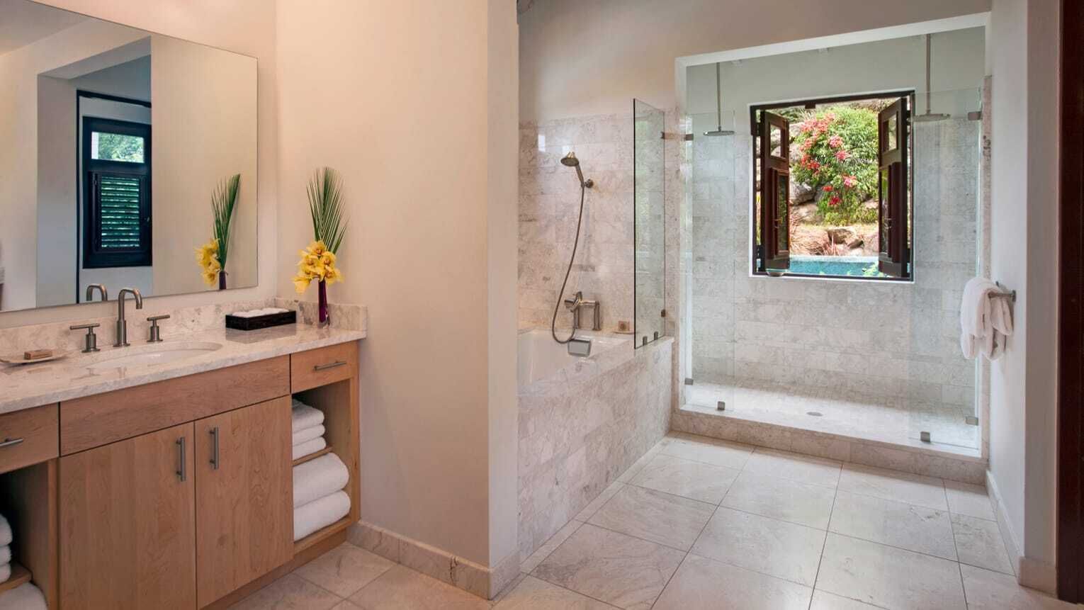 A modern bathroom with a large mirror, two sinks, a shower, and a view of a garden through a window.
