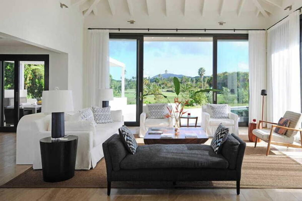 A modern living room with a sofa, two armchairs, a coffee table, and a TV stand. Large windows offer a view of a lush garden and hills.