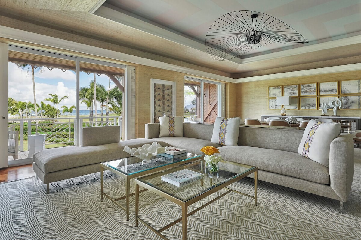 A spacious living room with a large sofa, a glass coffee table, and a view of the ocean. There is a dining area with a table and chairs, and a ceiling fan.