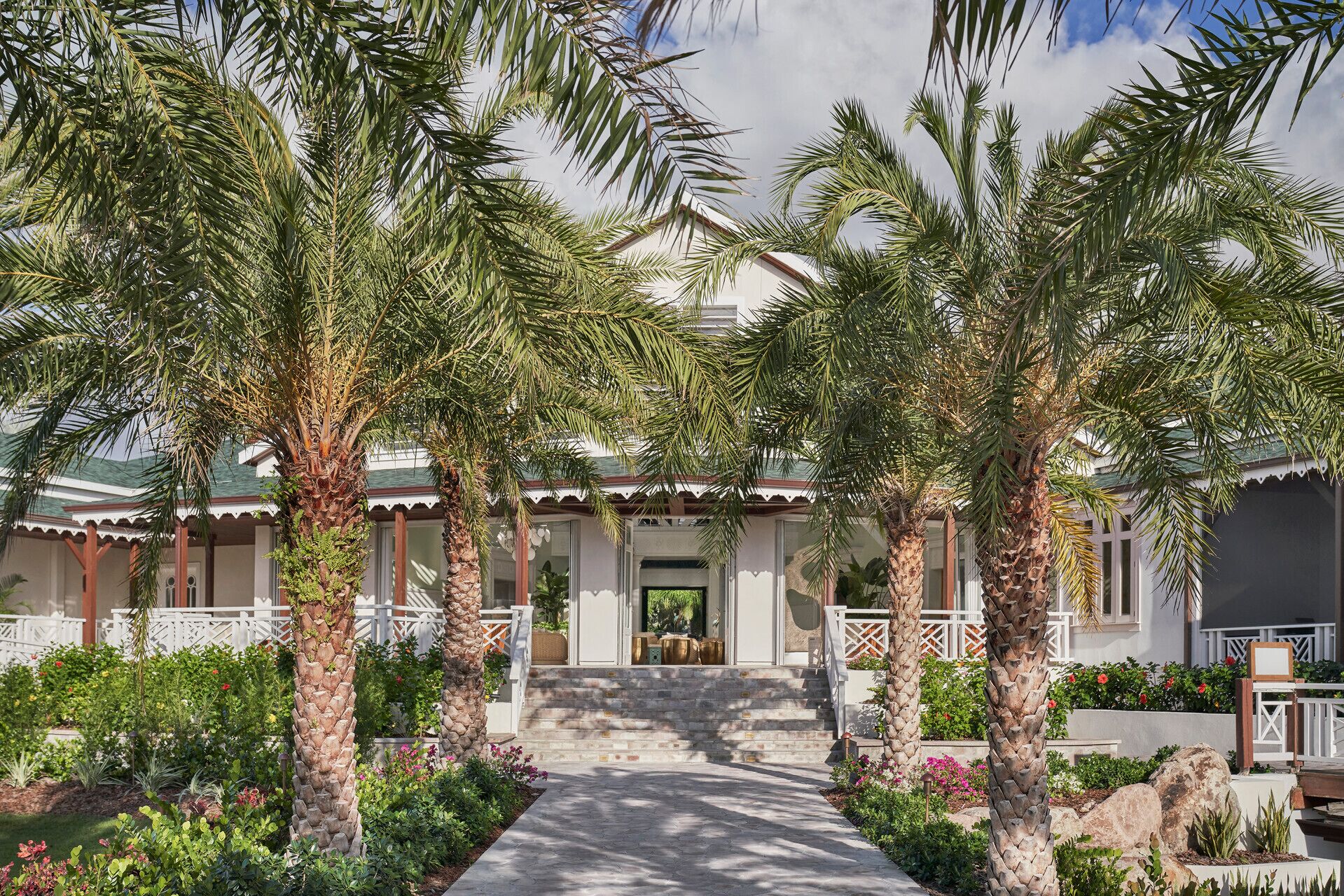 A house with a covered porch, palm trees, and a garden with various plants and flowers.