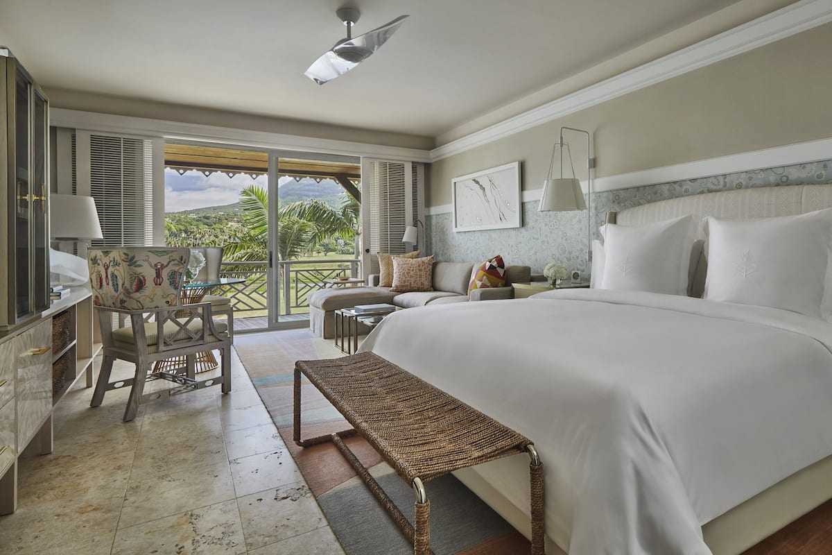 A spacious bedroom with a large bed, a balcony with a view of the outdoors, a seating area with a table, and a small side table with a lamp.