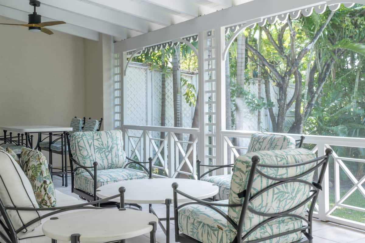 A covered outdoor seating area with metal chairs and tables, surrounded by a white railing, overlooking a garden with trees.