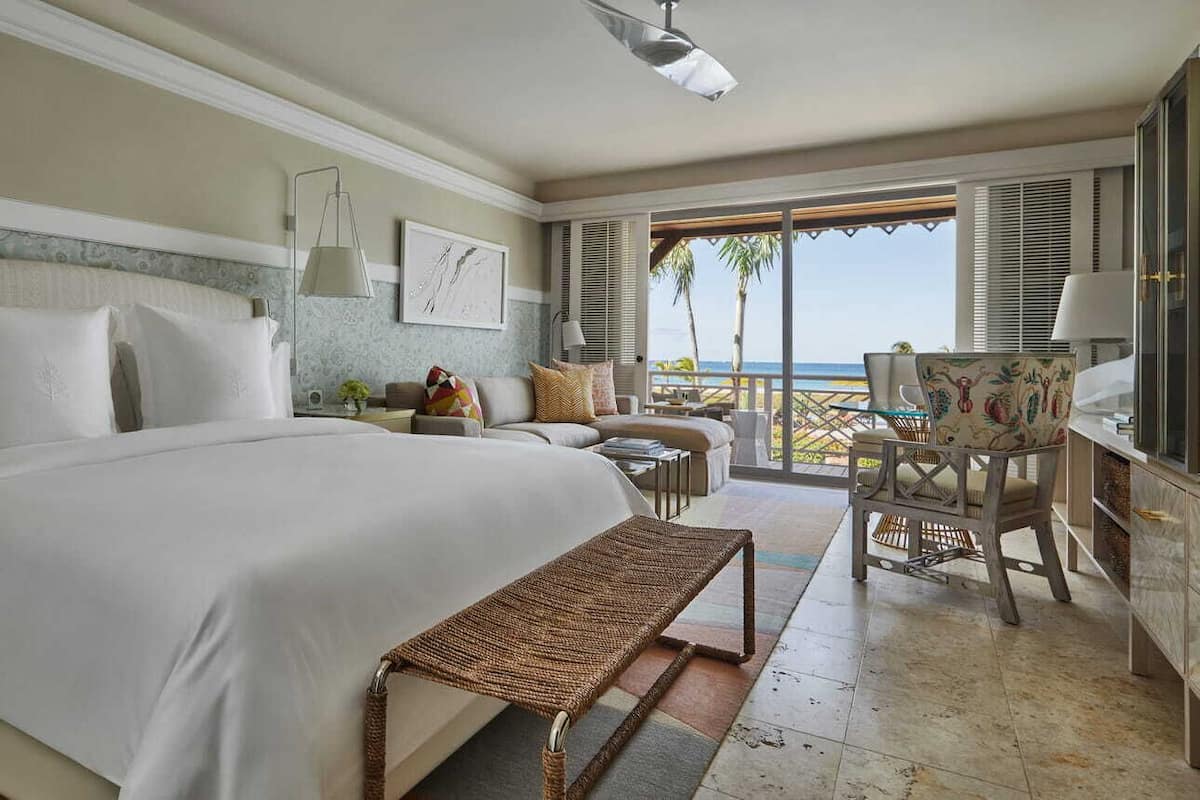 A hotel room with a large bed, a small table, a chair, and a view of the ocean through the window.