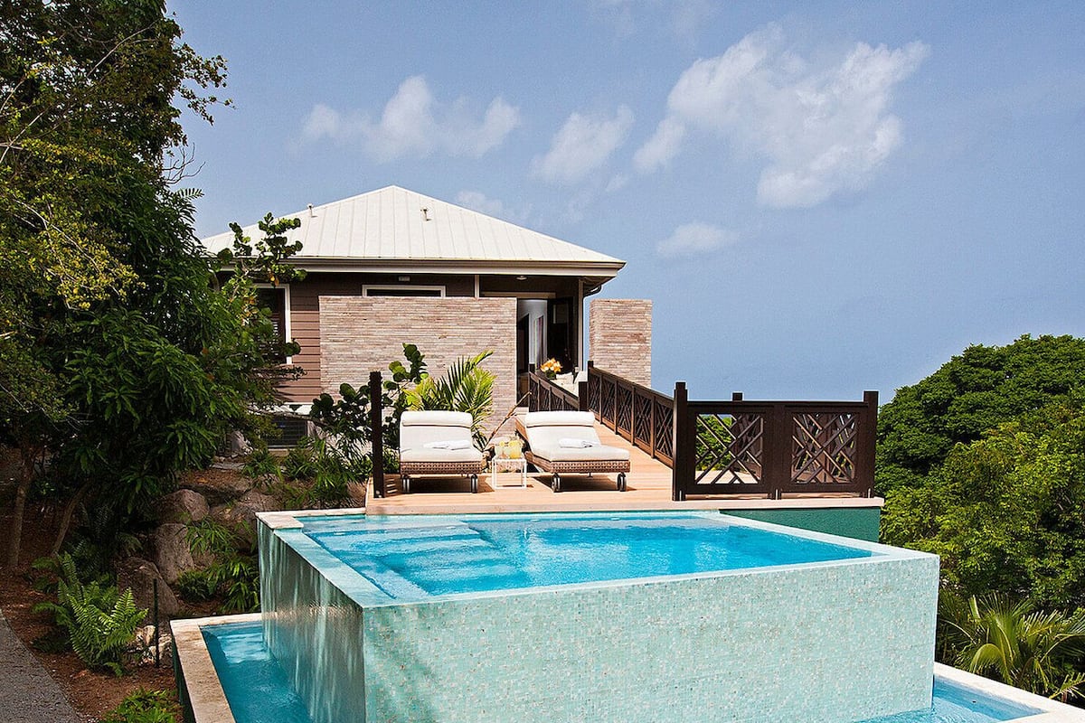 A rectangular swimming pool with two lounge chairs and a small table, surrounded by lush greenery and a building with a sloped roof.