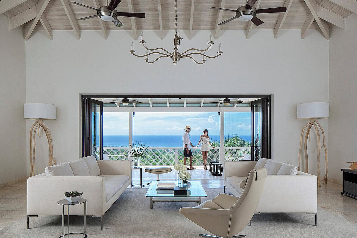 A spacious living room with two sofas, a coffee table, and a chandelier. There are ceiling fans and a large window offering a view of the ocean.