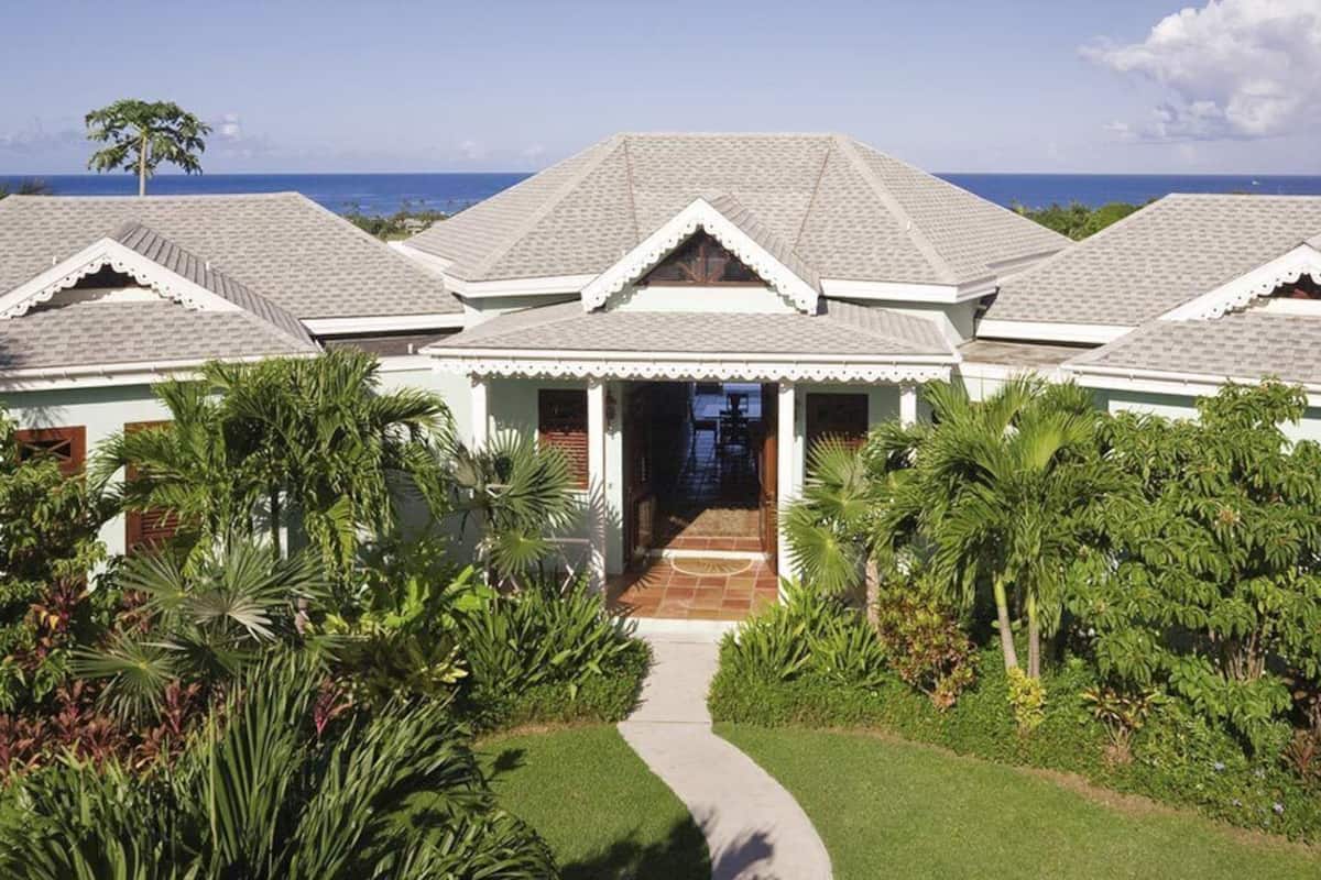 A two-story house with a tiled roof, surrounded by lush greenery and a clear view of the ocean.