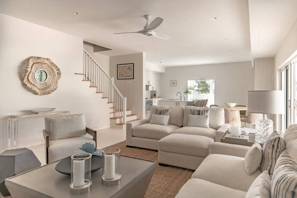 A modern living room with a neutral color palette, a staircase, and a kitchen in the background.
