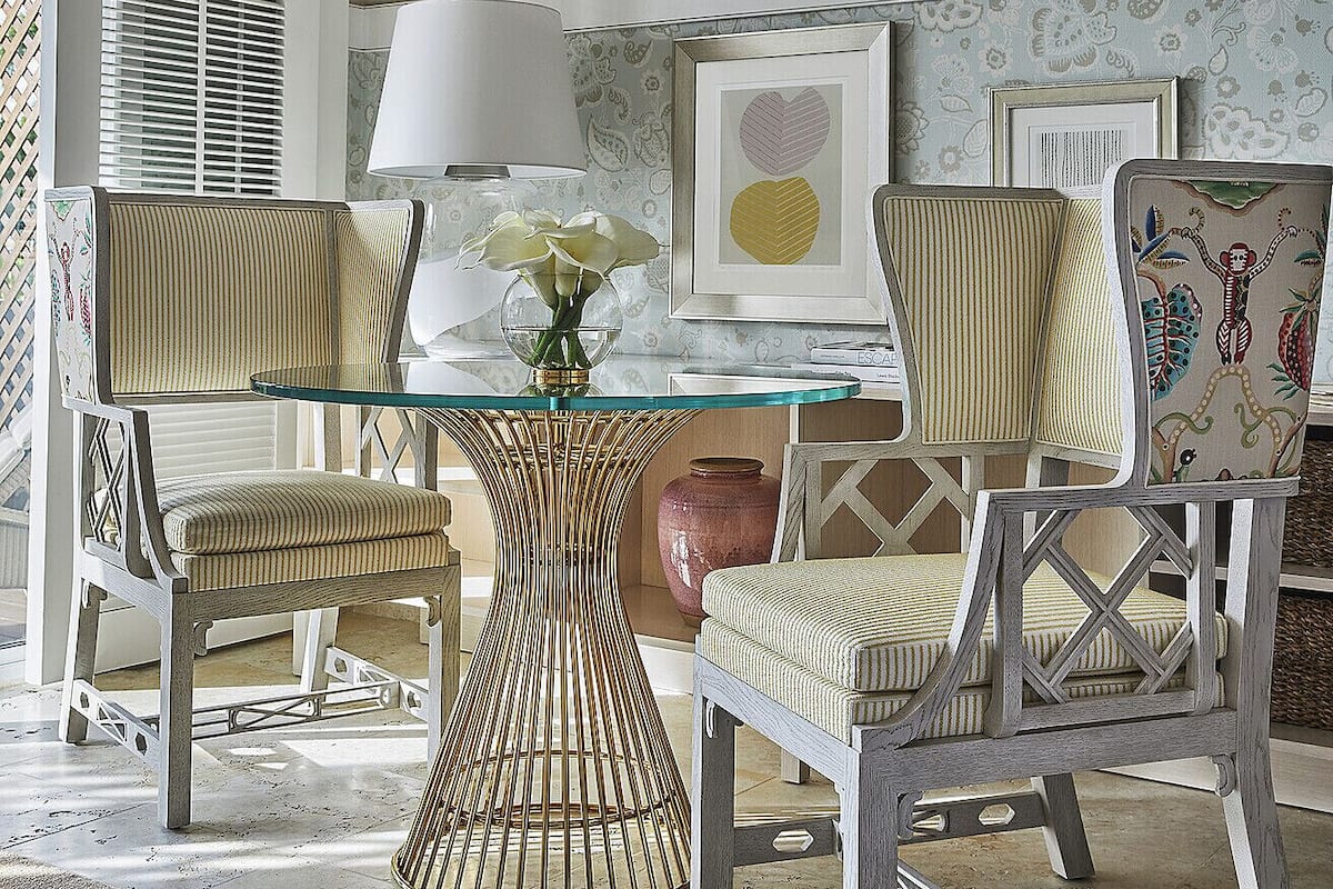 A dining area with a glass-top table, two upholstered chairs, a vase with flowers, and framed artwork on the wall.