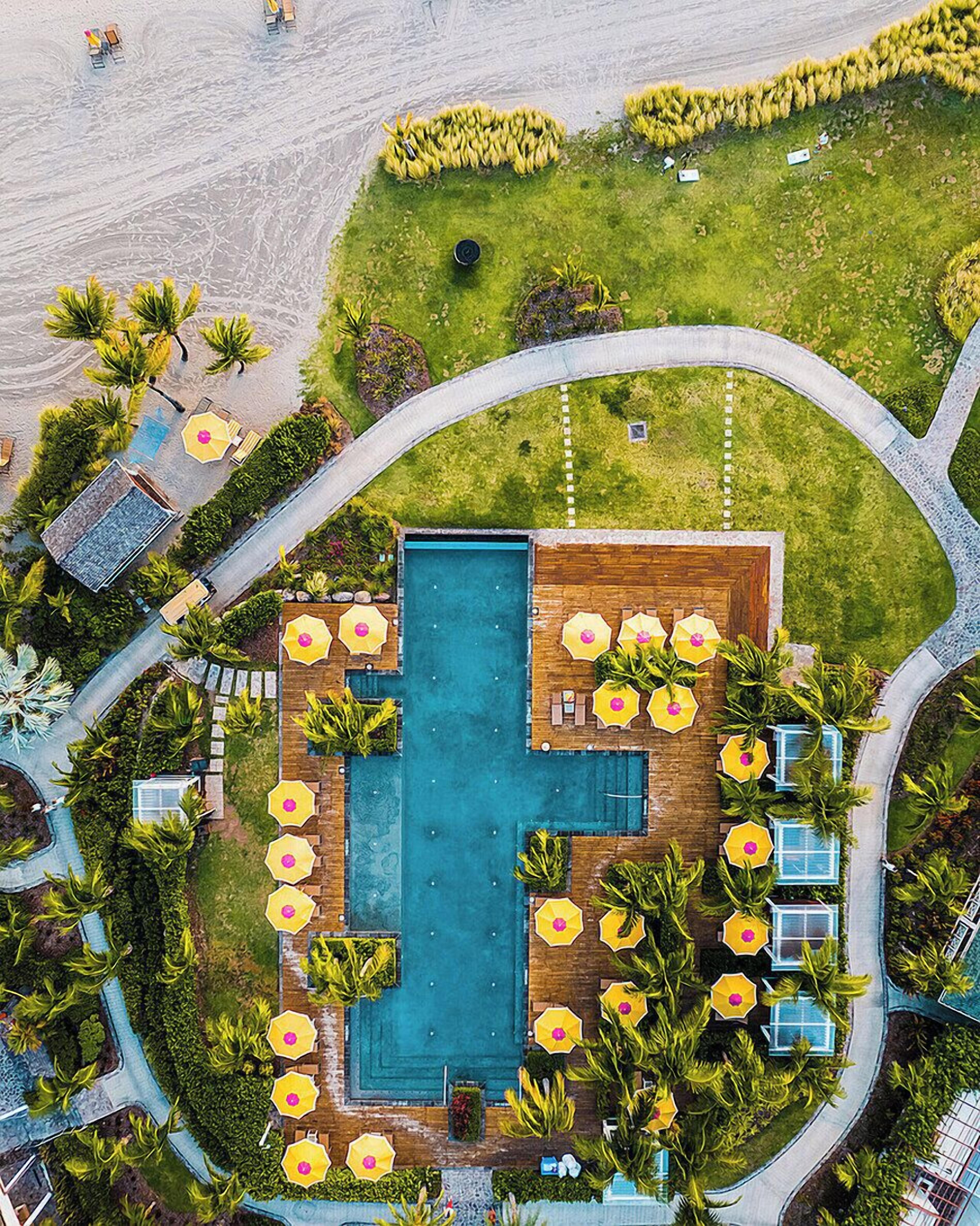 An aerial view of a resort with a central swimming pool, surrounded by lounge areas with umbrellas, and a well-maintained lawn.