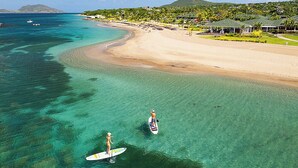 An aerial view of a beach with clear turquoise waters, a sandy shore, and a backdrop of lush greenery and hills. - Four Seasons Resort - Nevis (Jessups Village)