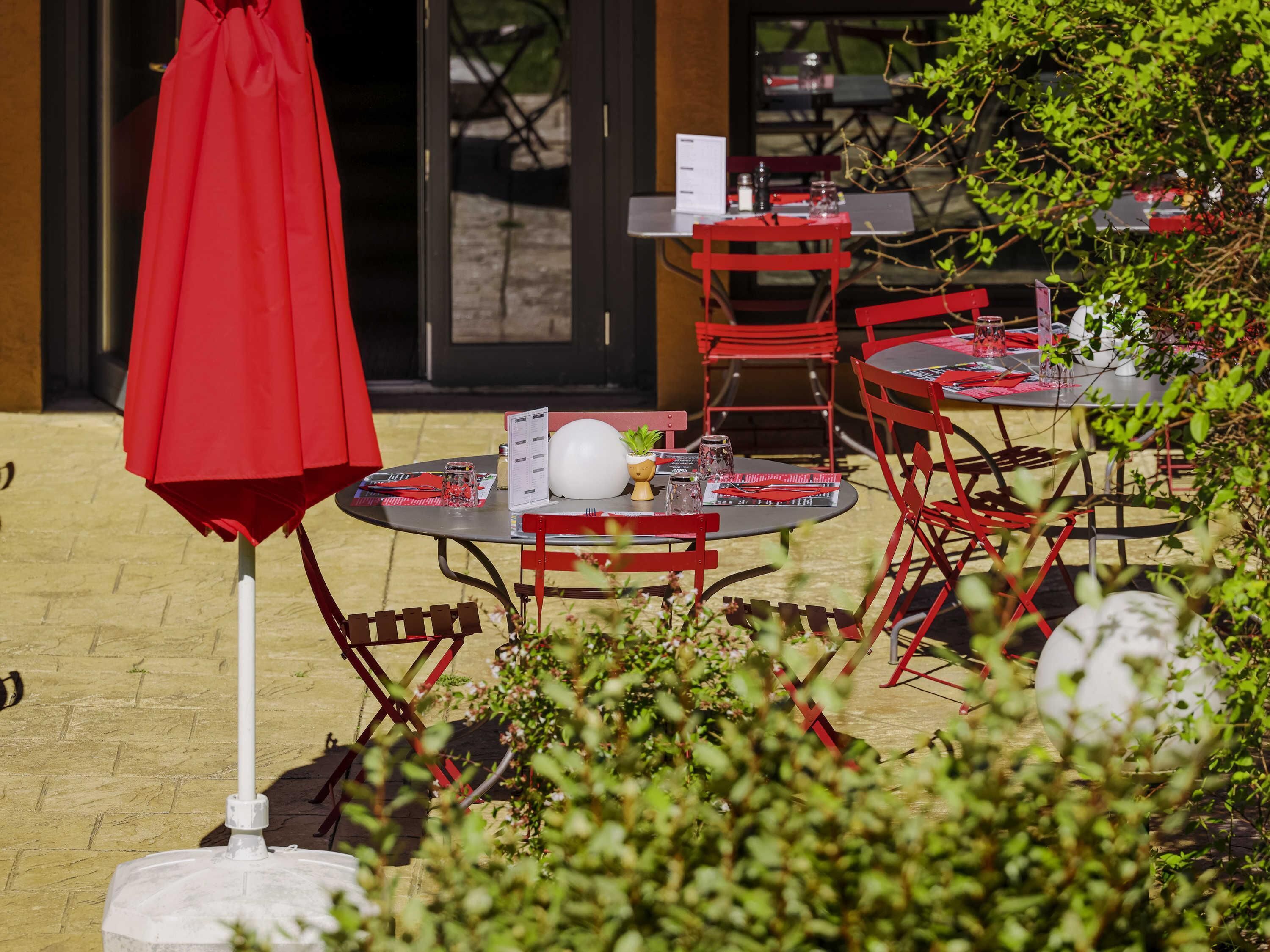 An outdoor seating area with red chairs and tables, a red umbrella, and a menu stand.