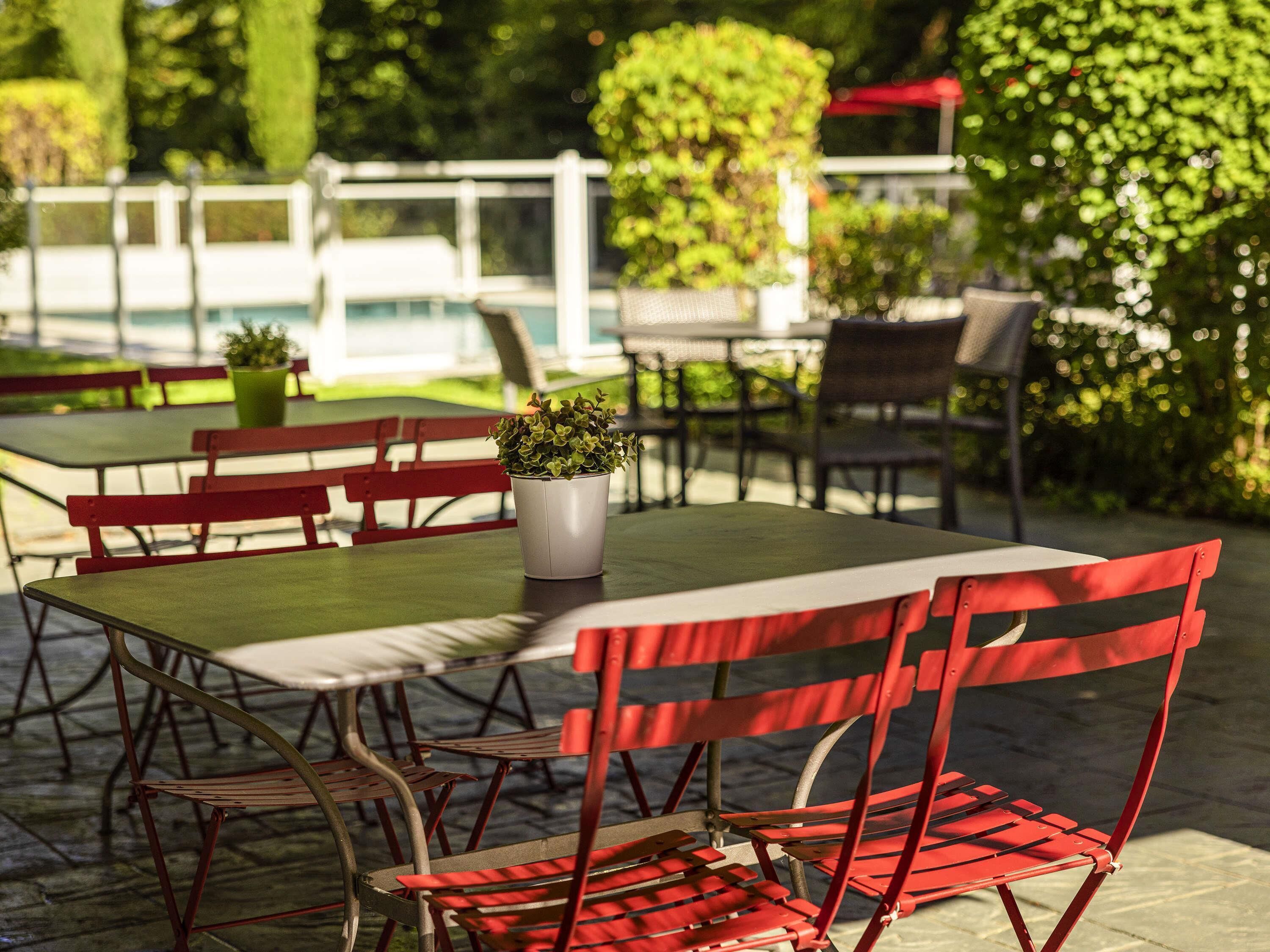 An outdoor seating area with red chairs and tables, a potted plant, and a white fence in the background.