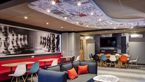 A modern lounge area with a curved ceiling, a large wall mural, and a variety of seating options including sofas, chairs, and tables. - ibis Lyon Nord (Dardilly)