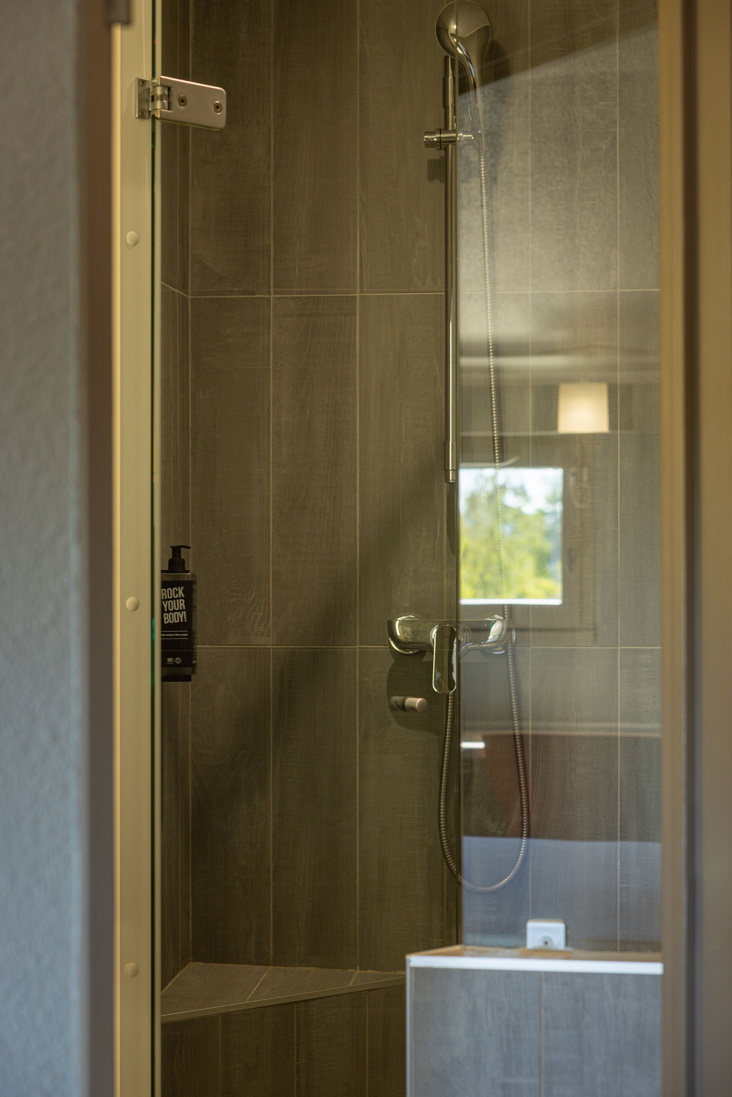 Ducha con mampara de vidrio, cabezal de ducha de mano y otro fijo, dispensador de jabón y una ventana pequeña que refleja follaje verde.