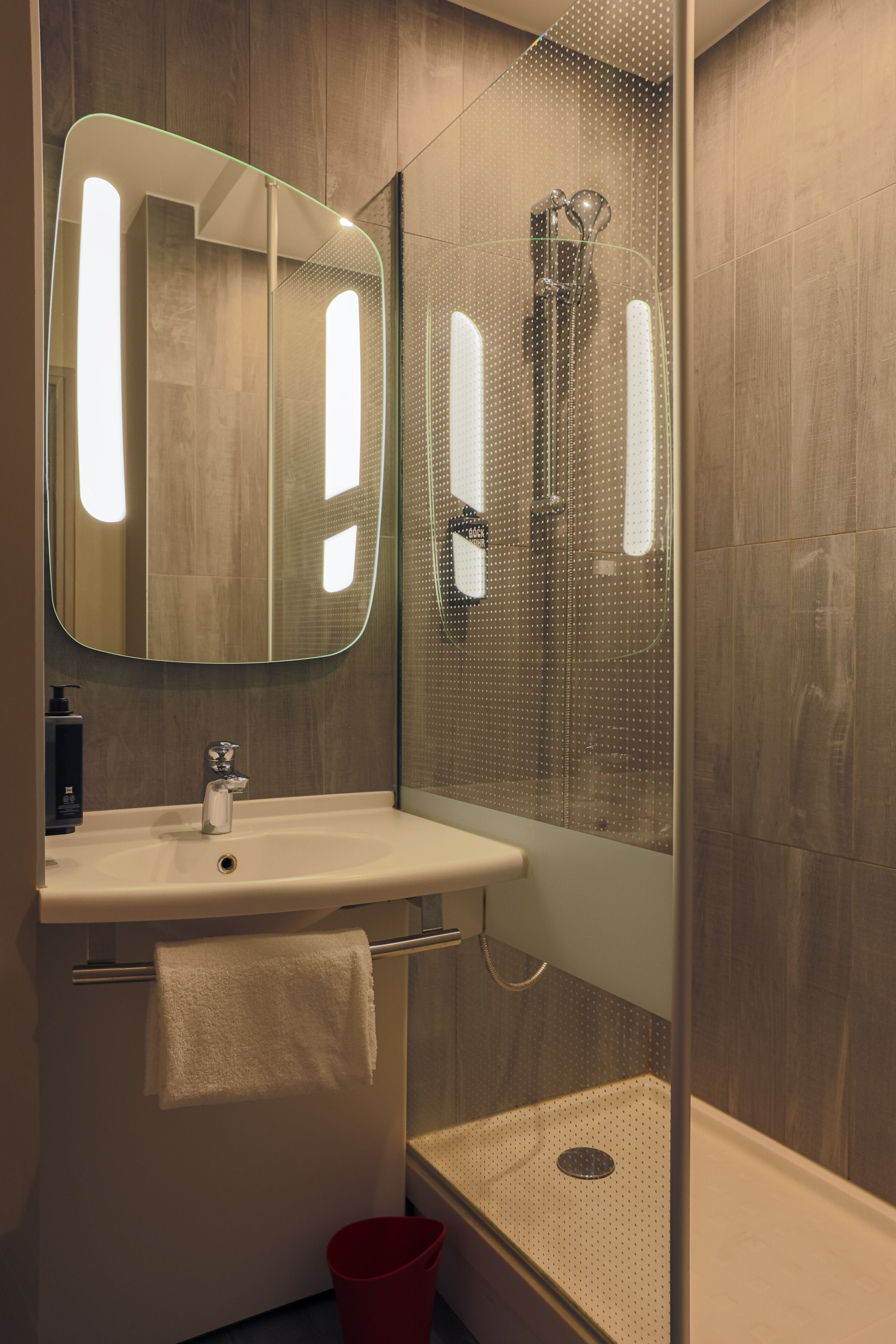 Une salle de bain moderne avec une douche à parois de verre, un lavabo blanc avec une serviette et un seau rouge.