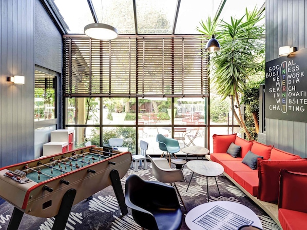 A modern indoor space with a foosball table, seating area, and a large window with blinds. - ibis Lyon Nord (Dardilly)