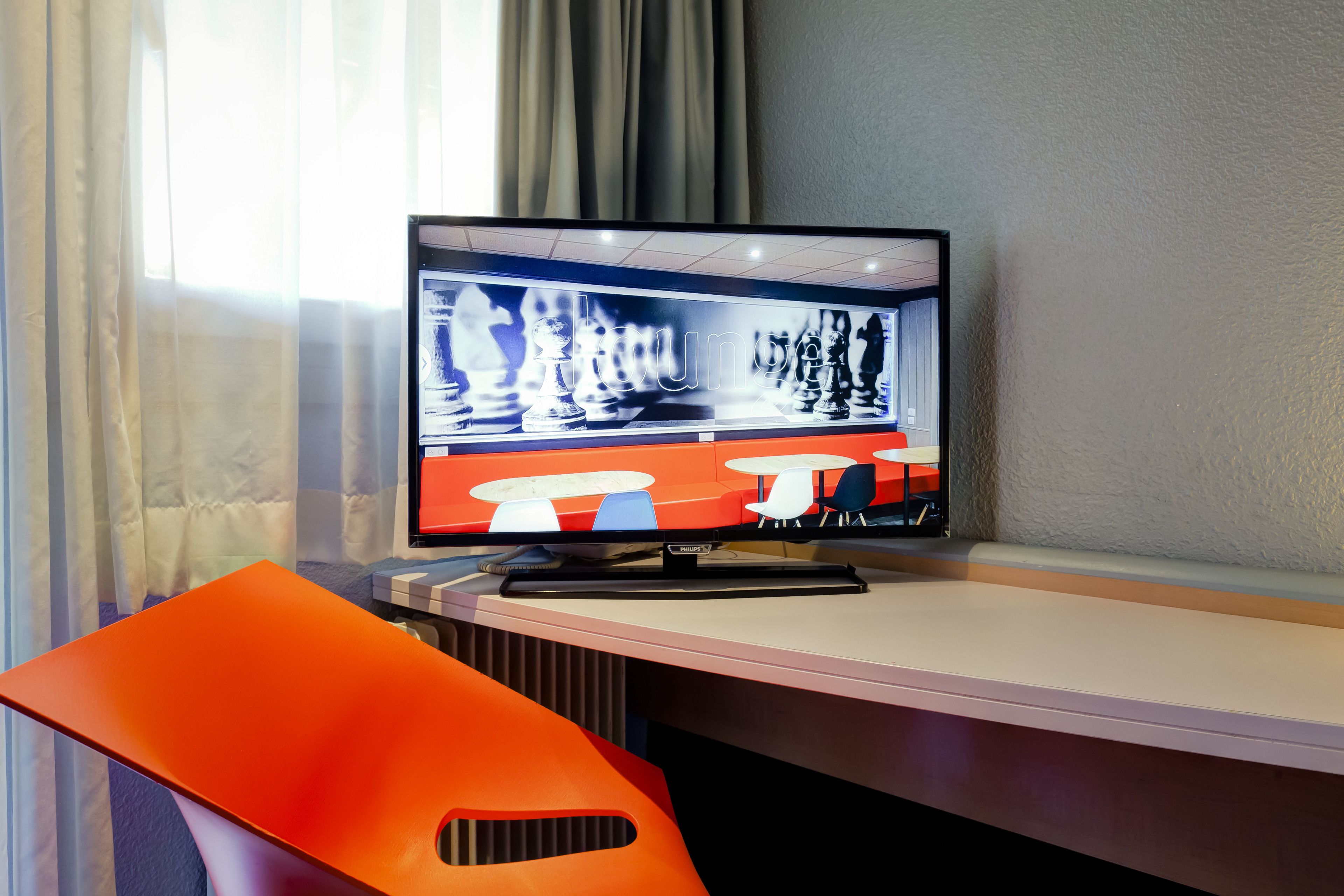 A modern room with a large flat-screen TV displaying a chess game, a bright orange chair, and a window with curtains.