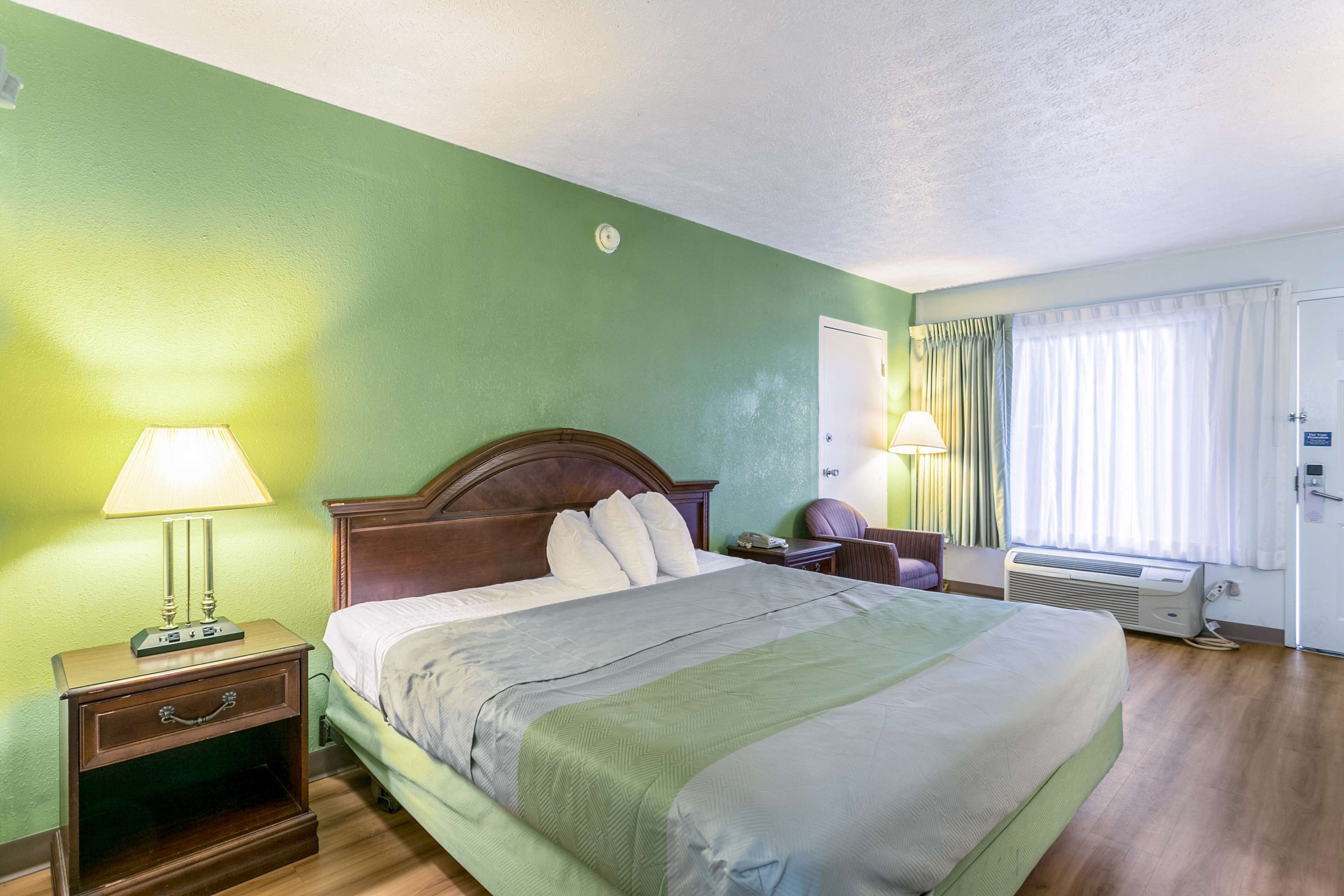 A hotel room with a bed, nightstand, lamp, chair, and air conditioning unit.