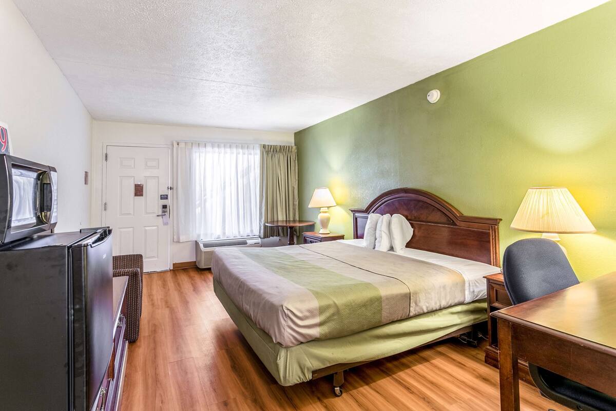 Quarto de hotel com cama, escrivaninha, cadeira, televisão e luminária.