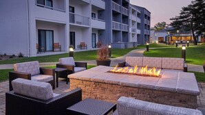 A modern outdoor seating area with a fire pit, wicker furniture, and a building with balconies in the background.