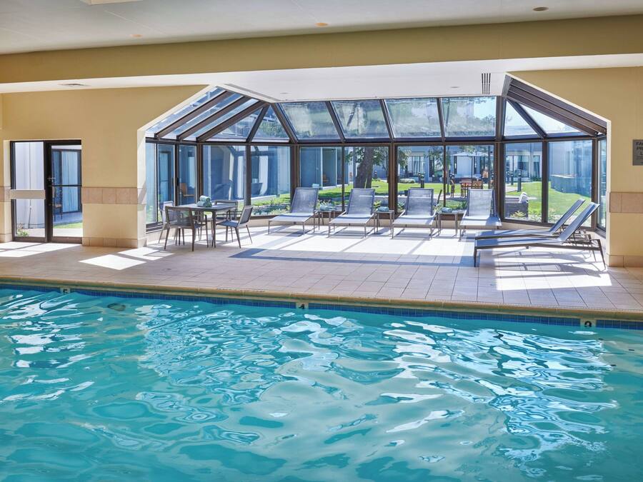 An indoor swimming pool with clear glass walls, lounge chairs, and a view of the outside garden.