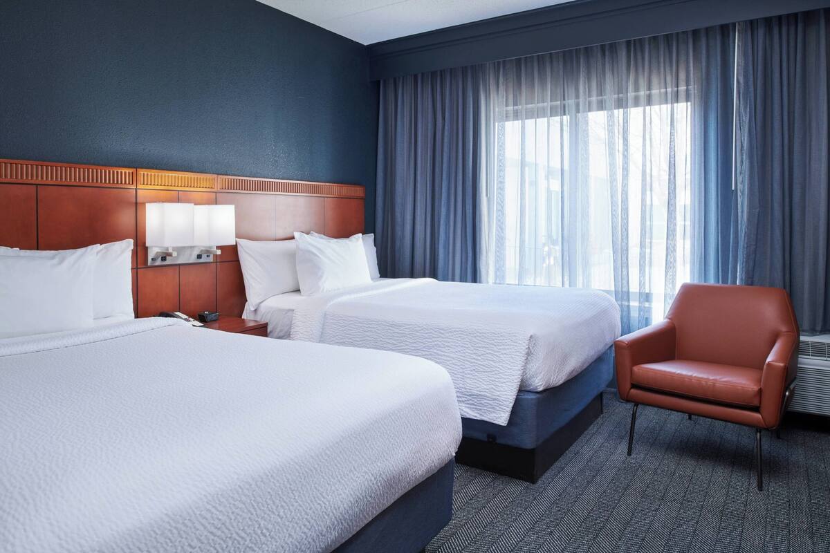 A hotel room with two beds, a wooden headboard, a chair, and a large window with curtains.