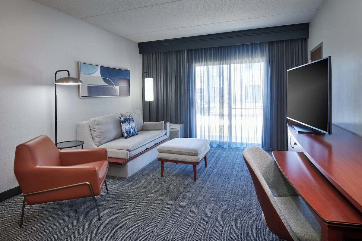 A hotel room with a sofa, armchair, ottoman, TV, and a large window with curtains.